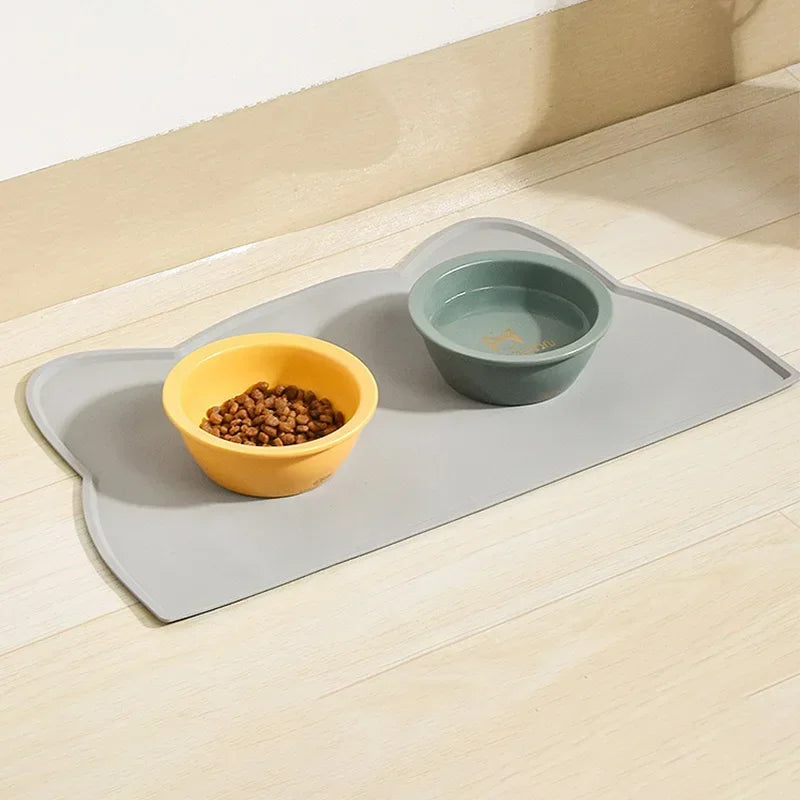 Gray silicone pet feeding mat with yellow food bowl and green water bowl on wood floor